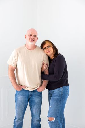 A couple wearing casual clothing stands together against a white wall, with one person leaning on the other.