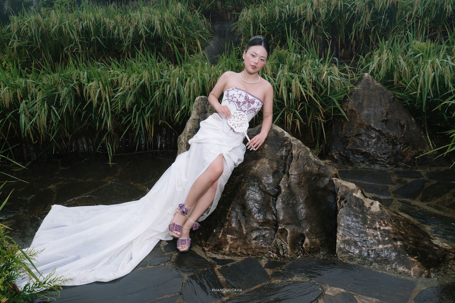 Bride in strapless white embroidered gown with high slit poses on rocks surrounded by green reeds.