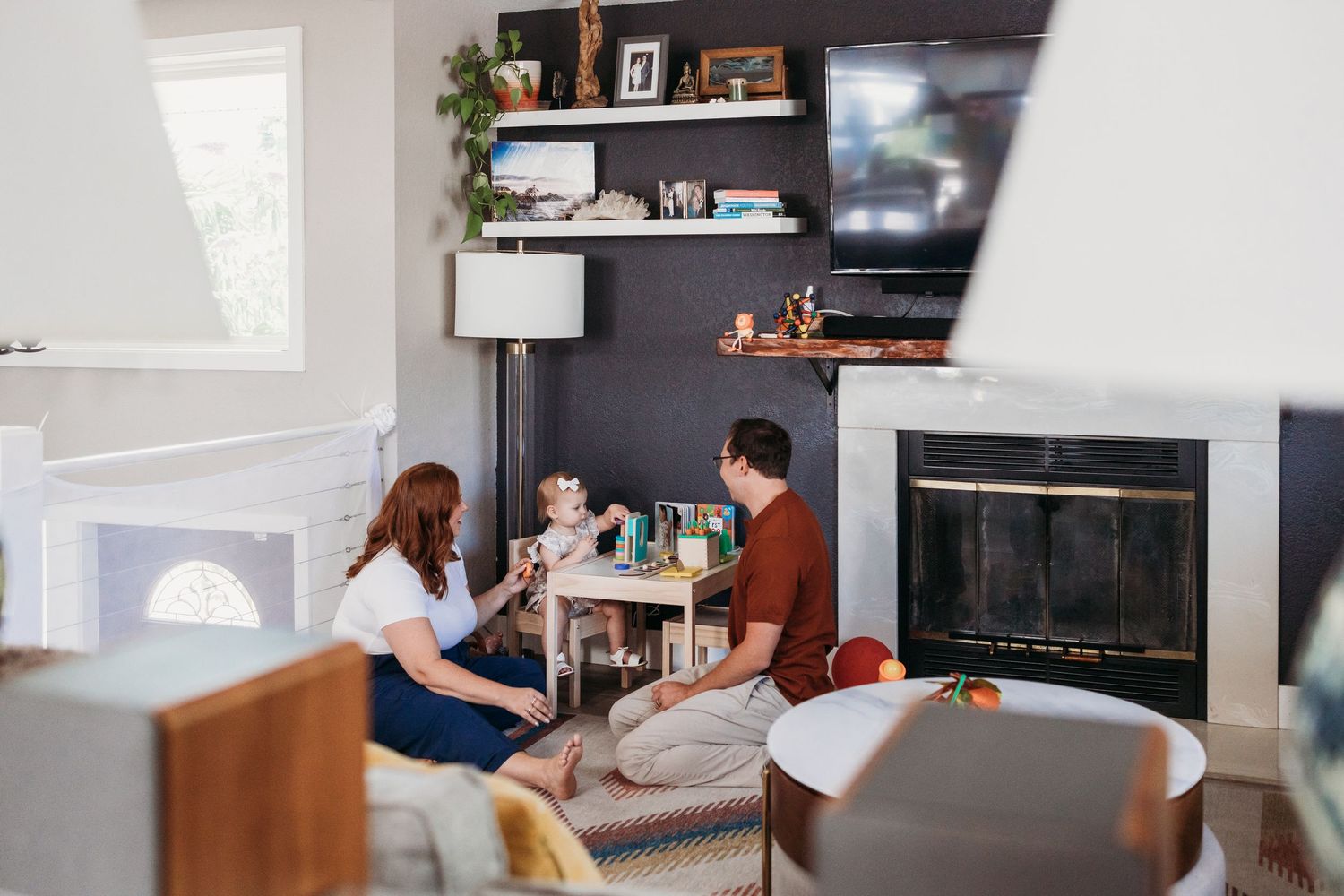 Two people sit in a modern living room with dark accent walls, floating shelves, and a fireplace.