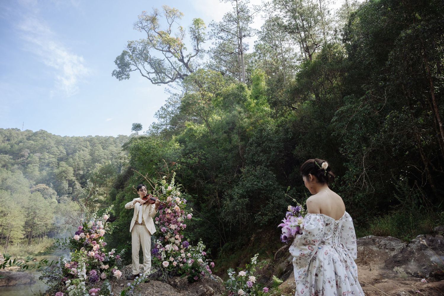 Bride in floral gown and groom pose on rocky cliff surrounded by lush forest and purple flowers.