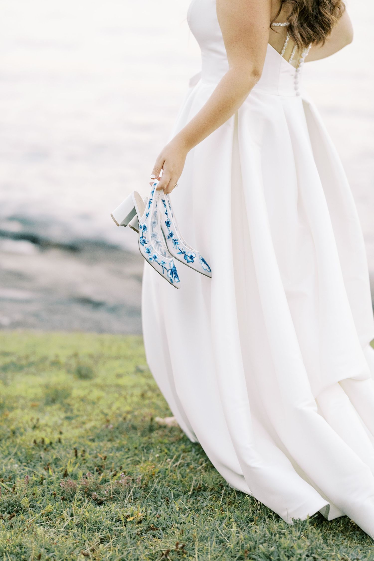 Close-up detail of blue floral embroidered wedding shoes peeking out under flowing white dress on grassy lawn.