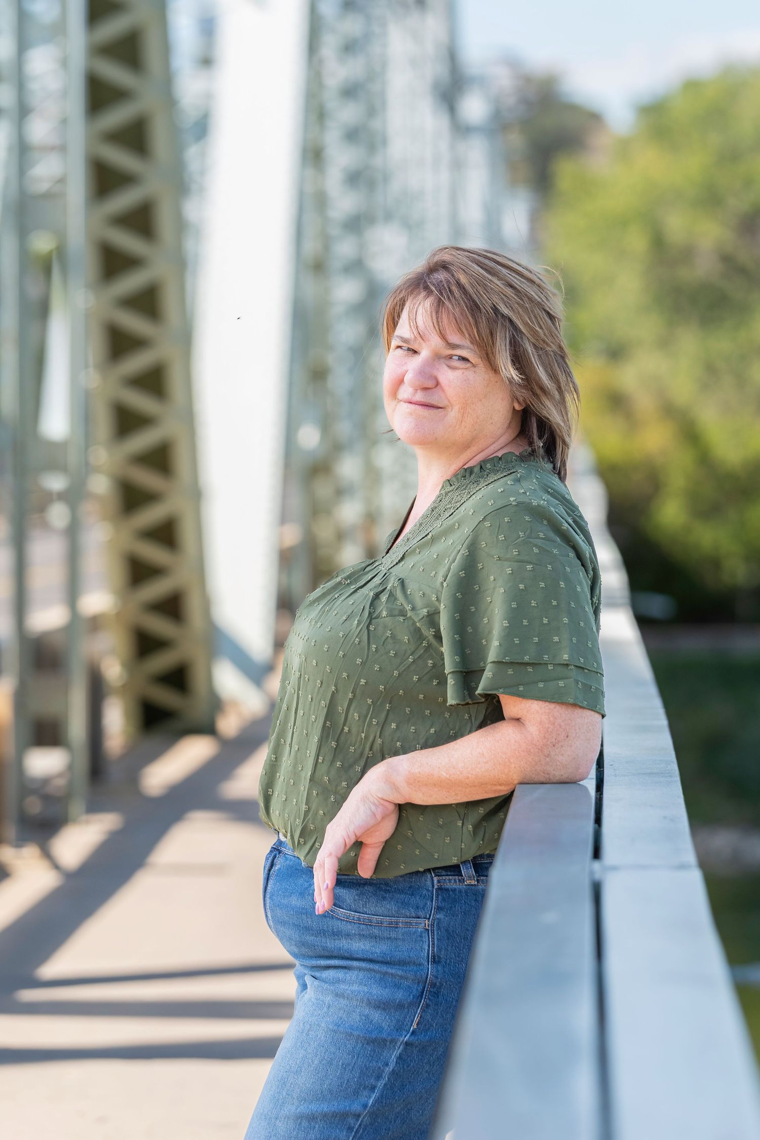 Woman in olive green top and jeans smiles in outdoor photo series against metal bridge structure.