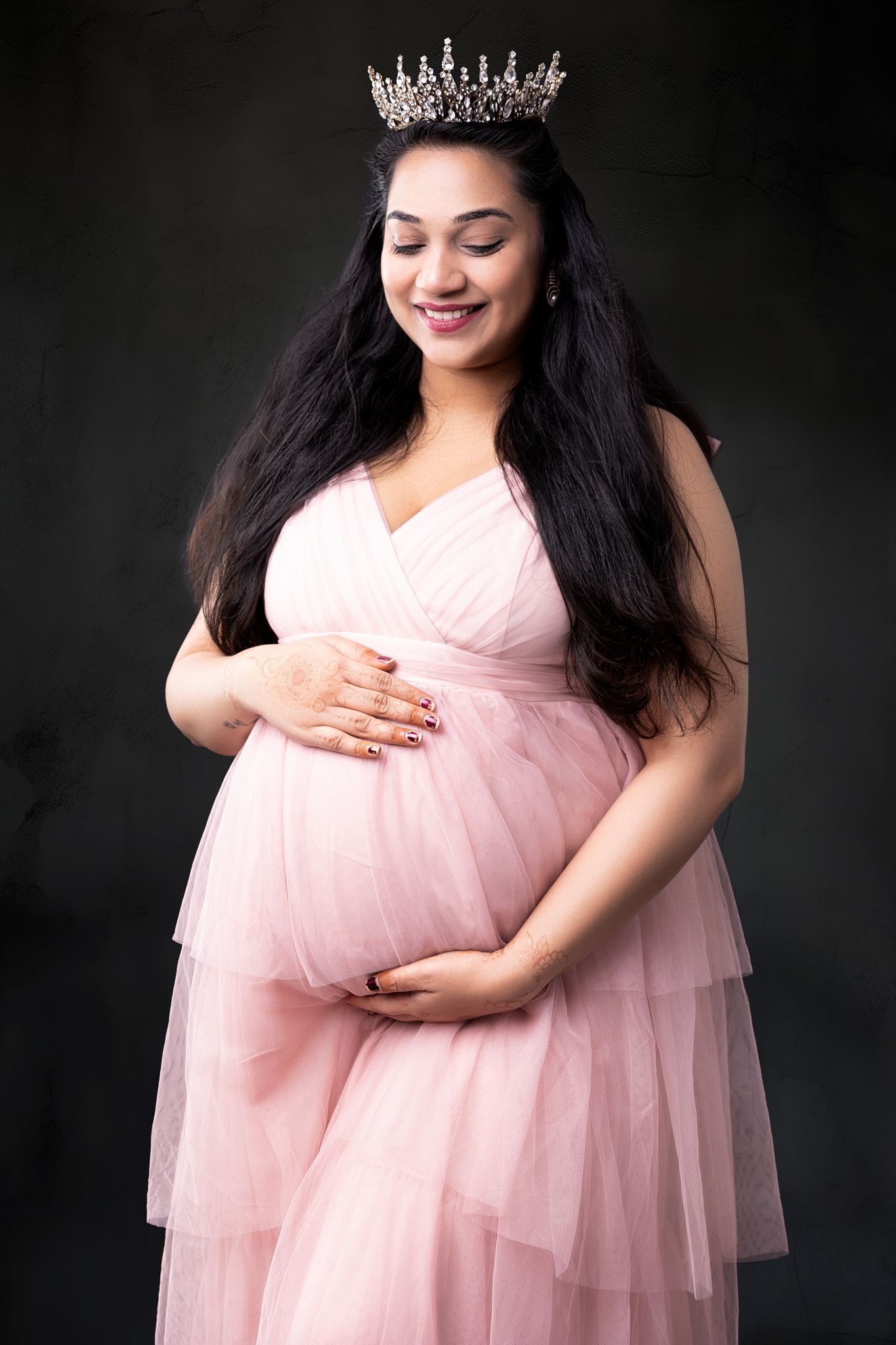 Maternity photo session showing a woman in a flowing pink tulle dress against a dark background.