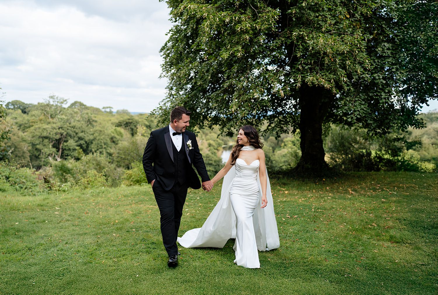 Bride and groom walk hand in hand through a lush green hillside garden on their wedding day.