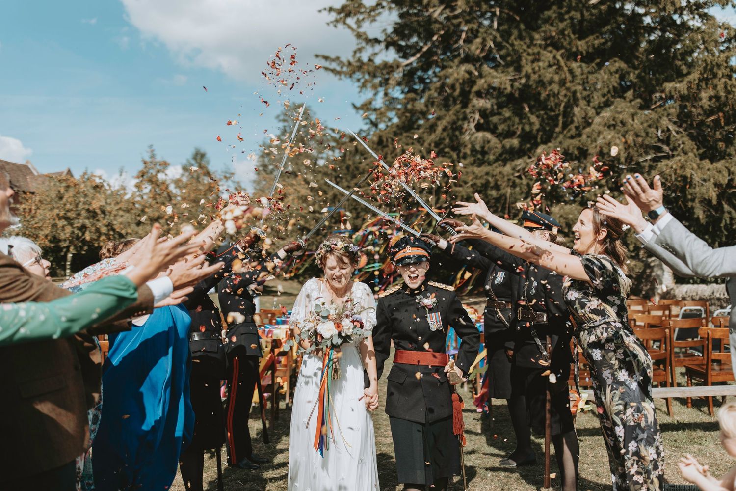 Wedding guests throw white flower petals during a joyful outdoor celebration on a sunny autumn day.