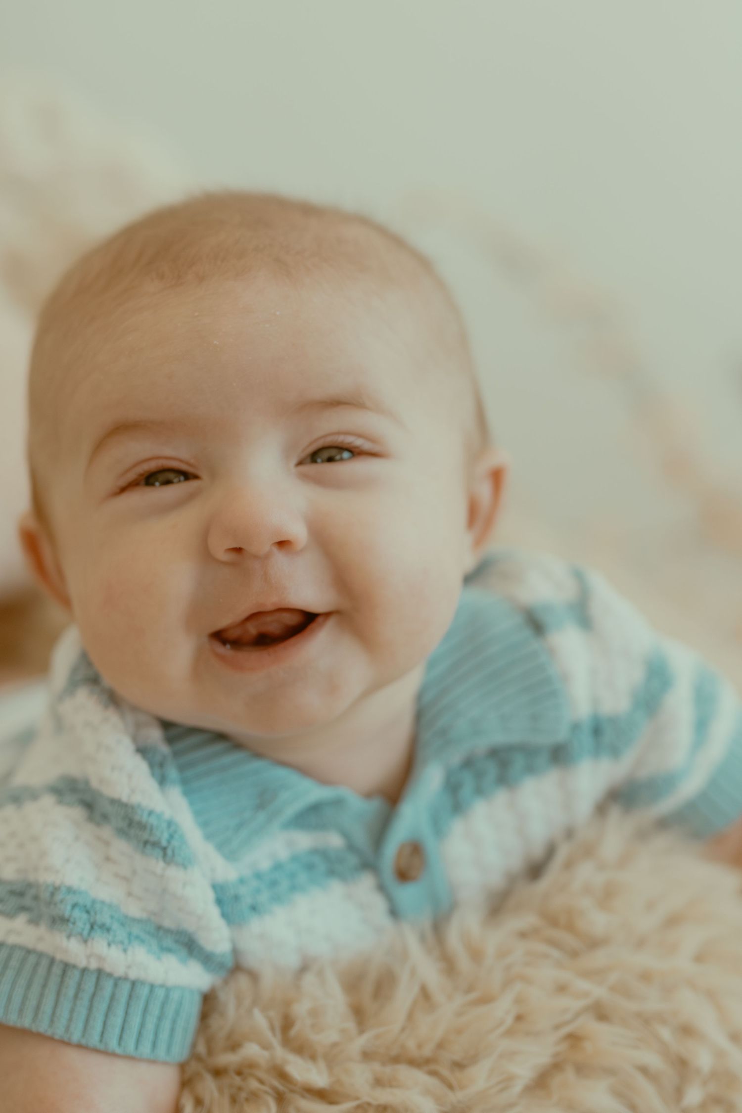 Sweet baby wearing light blue cardigan smiles joyfully while laying on soft beige blanket.