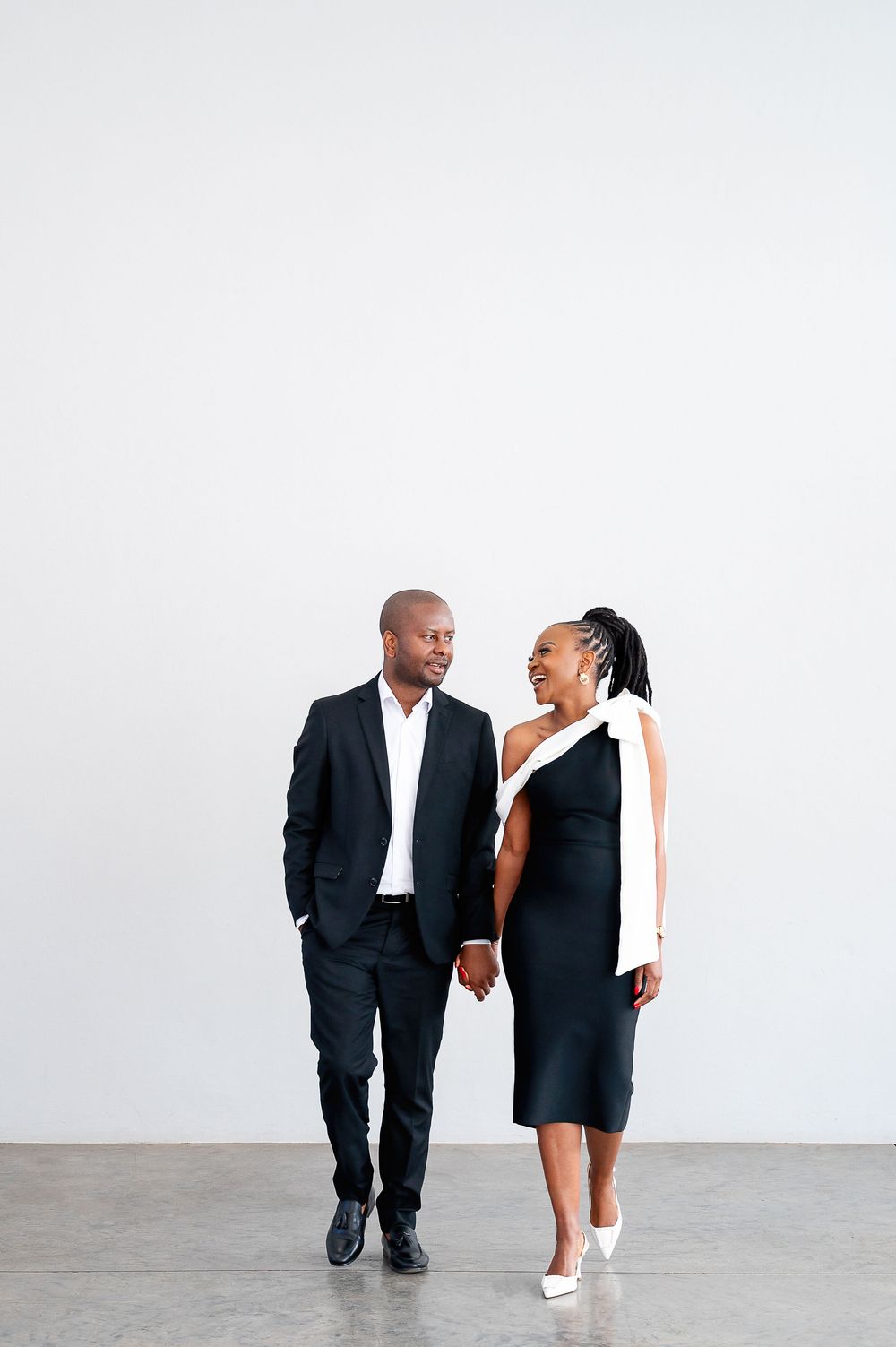 Couple in matching black and white outfits share joyful laughter and hold hands in front of a white wall.