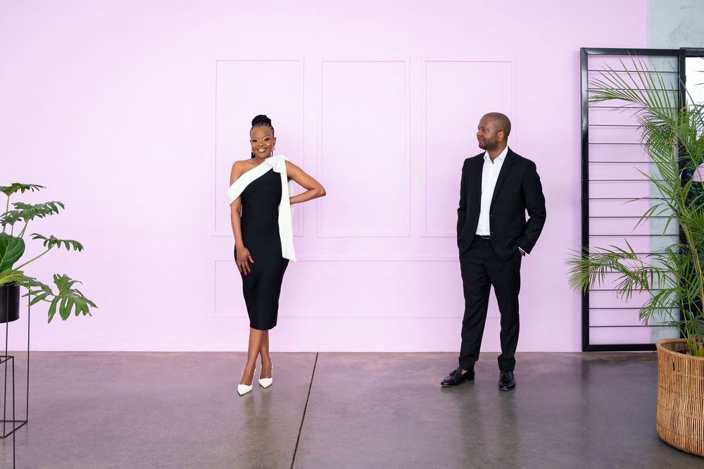 People in formal attire stand against a white wall during what appears to be a professional photoshoot.