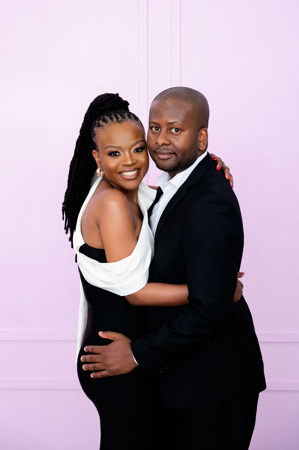 Romantic couple in formal black and white attire poses against pink wall for portrait.