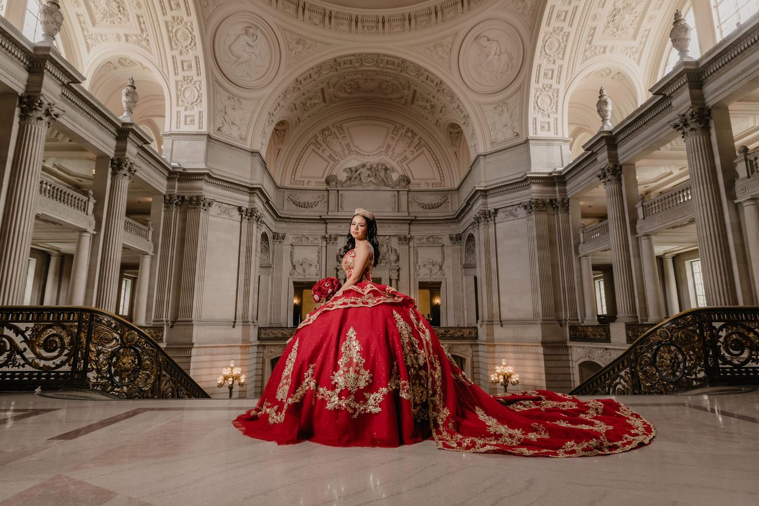 Model in red ballgown poses elegantly in ornate classical ballroom with white marble columns and grand architecture.