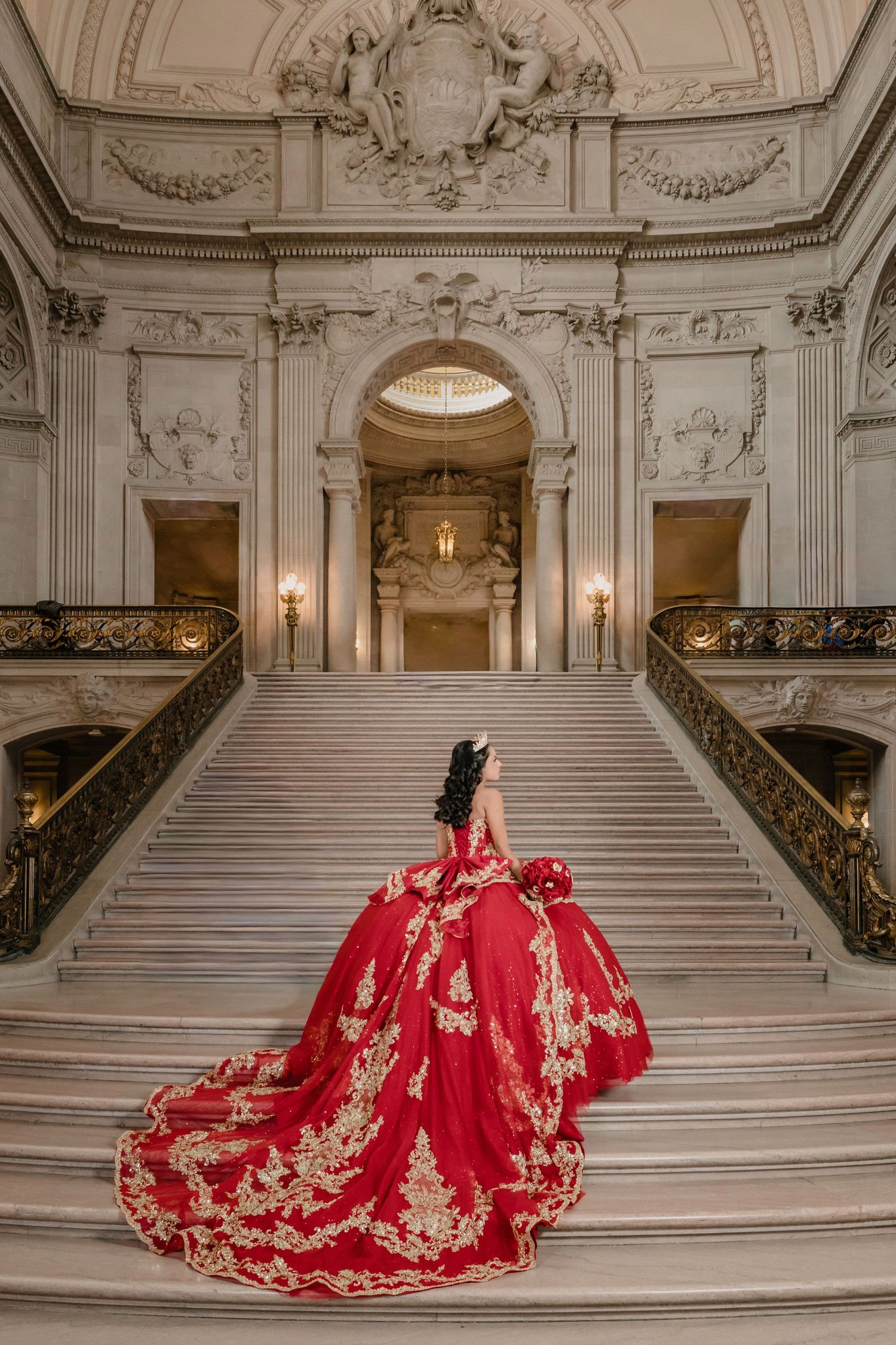 Quinceanera in ornate red and gold gown poses on grand marble staircase in luxurious palace interior.