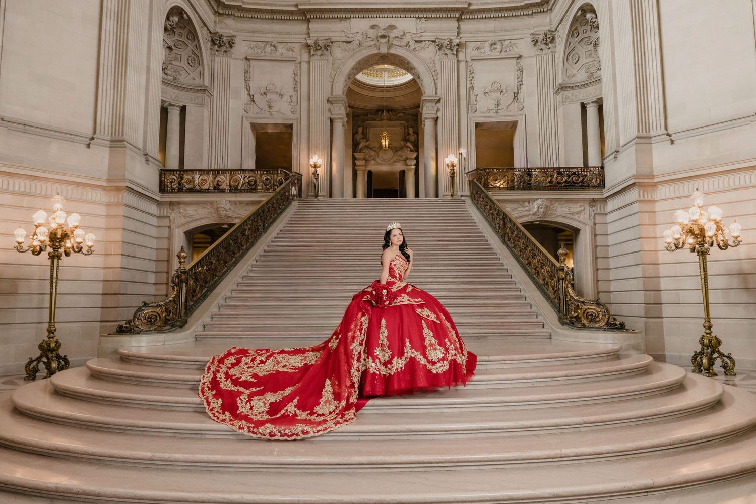 A person in a flowing red ballgown poses on a grand marble staircase in an ornate classical building interior.
