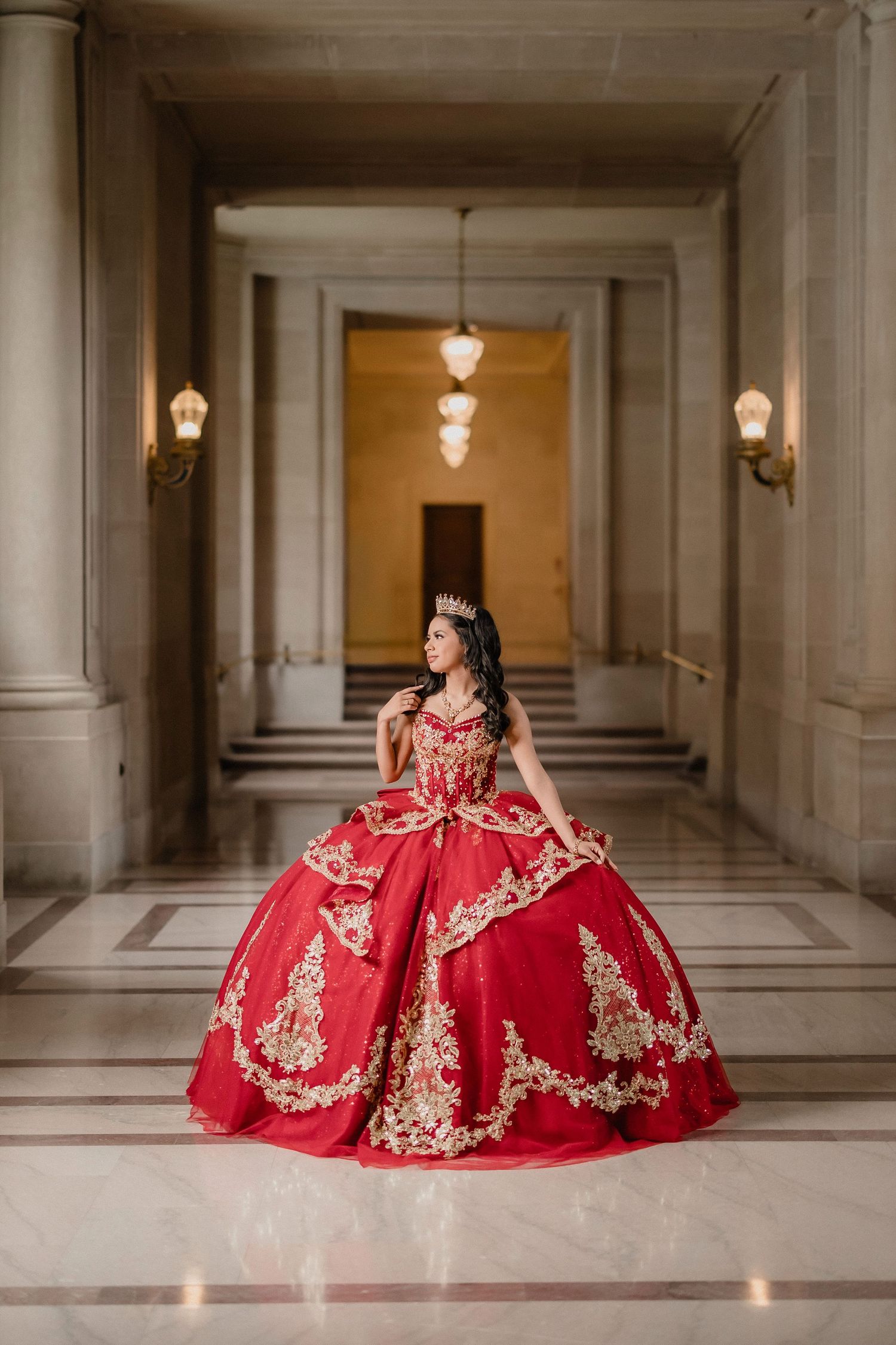 A stunning quinceañera photo session featuring an elegant red and gold ball gown in a grand marble hallway.