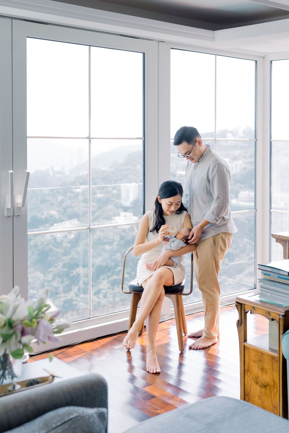 A couple stands by large windows in a modern apartment with mountain views while holding a small dog on their lap.