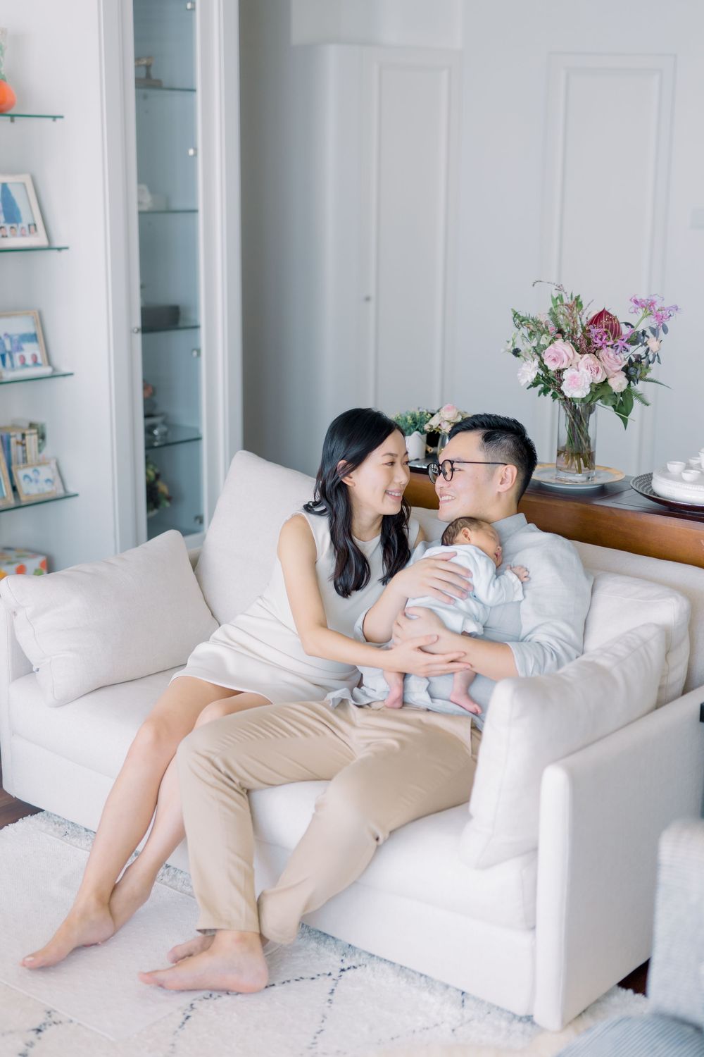 A couple shares an intimate moment on a white couch with their baby in a bright modern living room with floral decor.