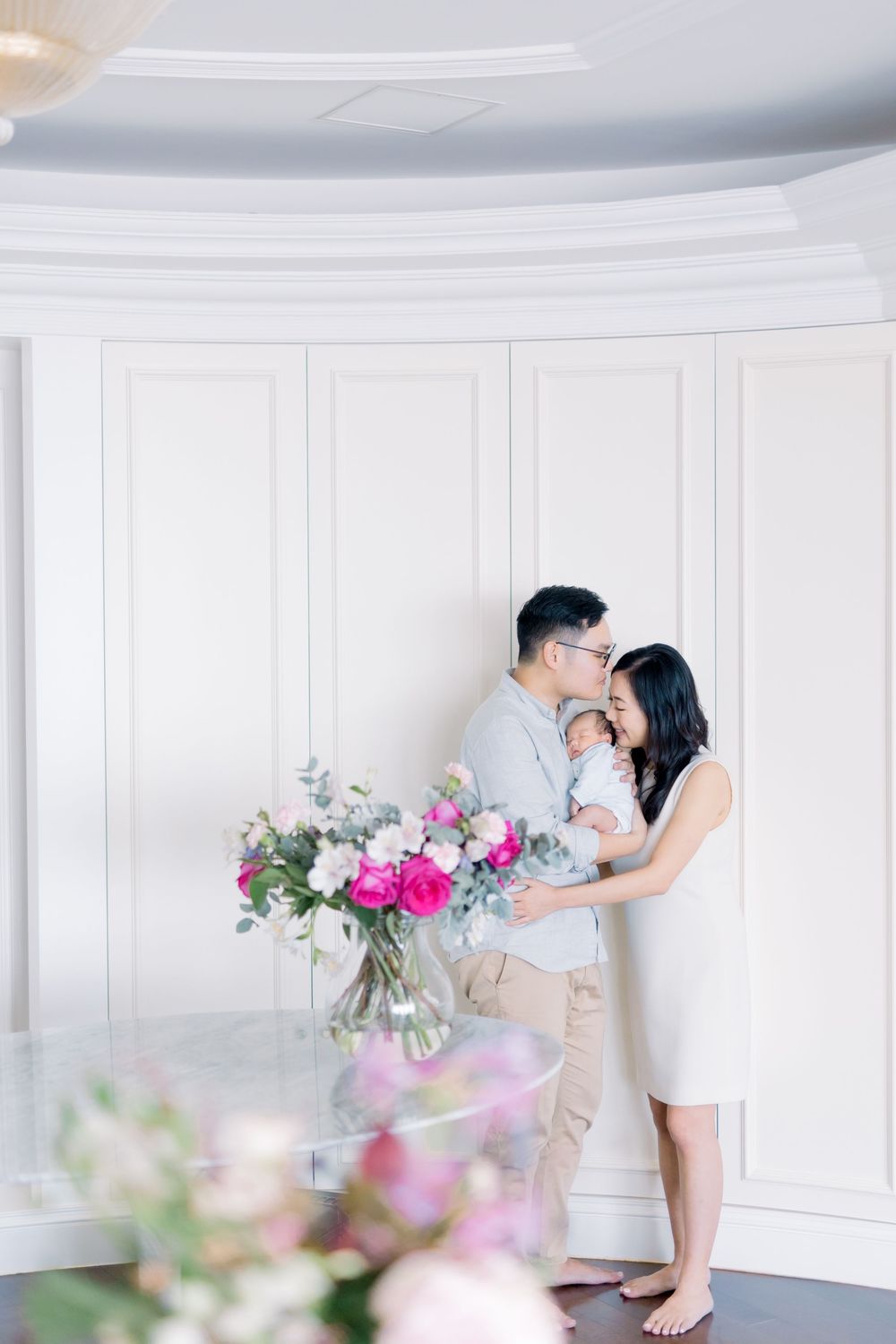 A romantic couple embraces in an elegant white room with soft lighting and pink floral arrangements in the foreground.