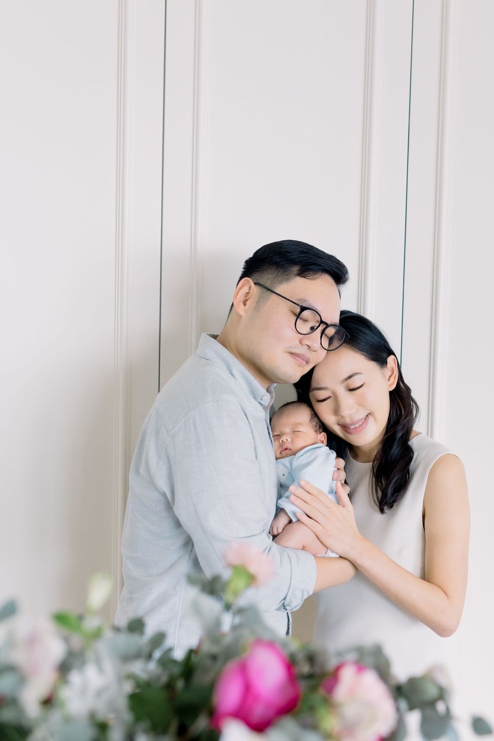 A couple in grey and white outfits share tender moments with their newborn baby against a white backdrop with pink flowers.