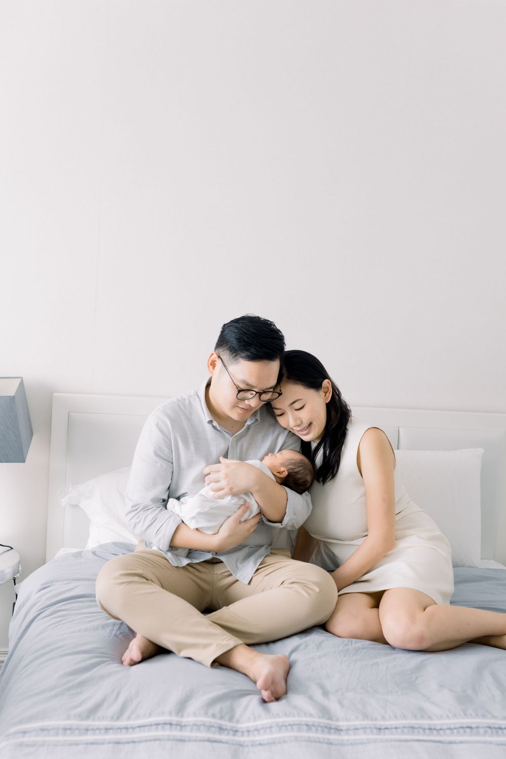 A couple lounges together intimately on a white bed wearing casual clothing in a bright minimalist setting.