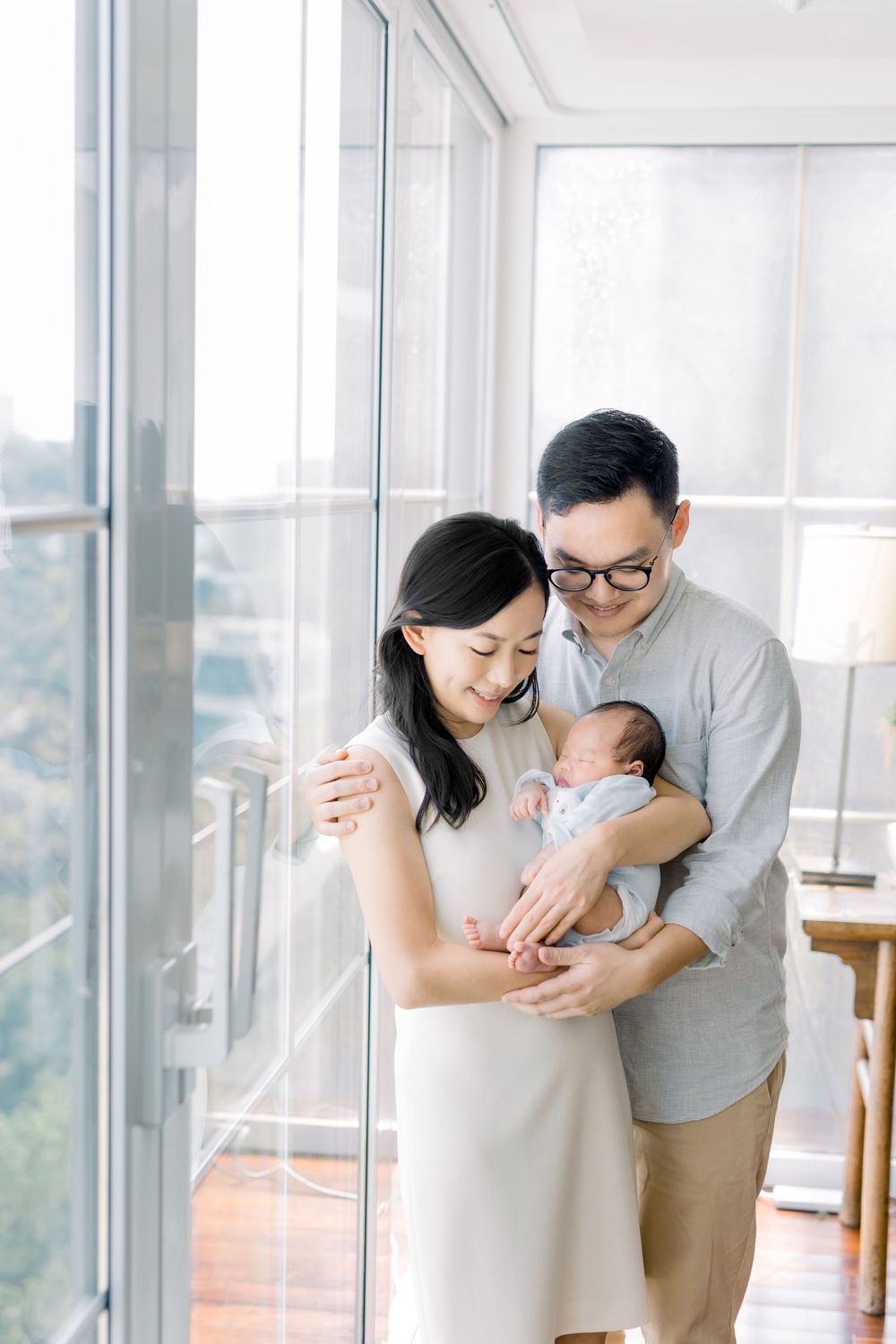 Young couple holding their newborn baby while standing by bright windows in a modern high-rise apartment.