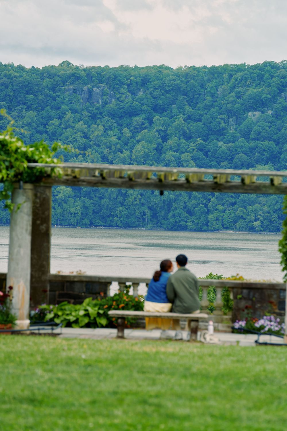 A scenic view of Lake Peekskill shows a distant shoreline with lush green forest and a grassy area in the foreground. Wave hill garden
