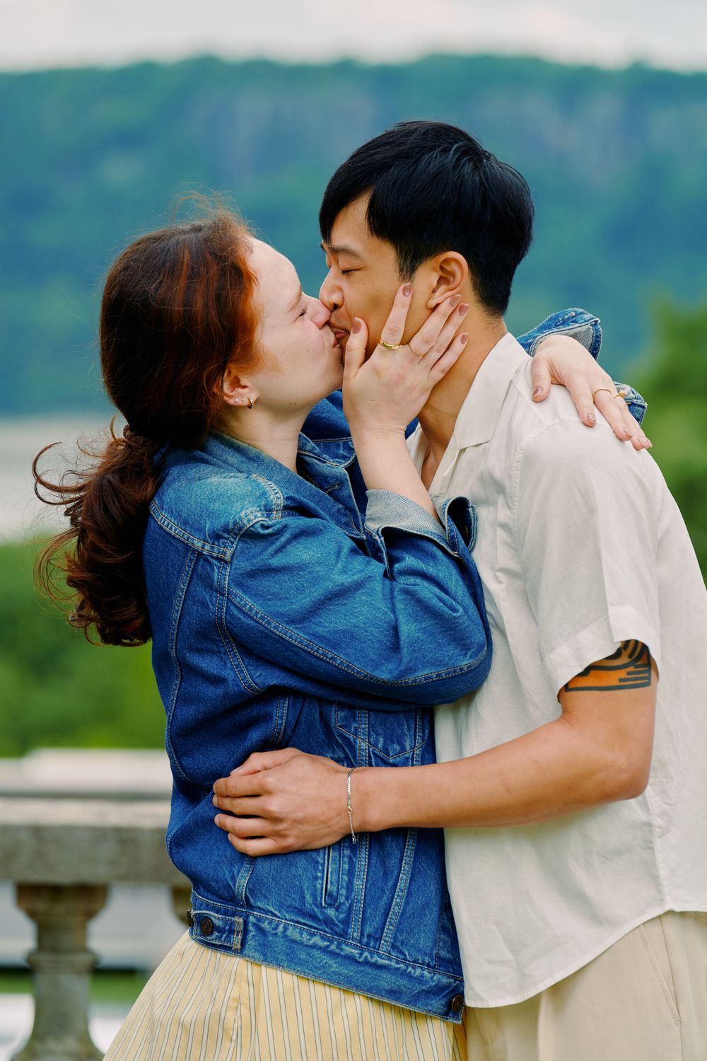 A romantic couple sharing a tender kiss outdoors, with one wearing a denim jacket and the other in a light colored shirt. Wave hill garden