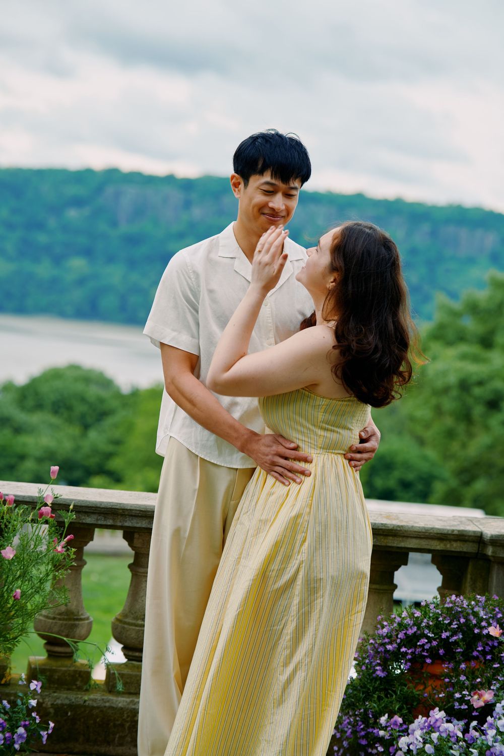 Romantic couple in coordinating light outfits embracing on a terrace with mountain views and purple flowers. Wave hill garden
