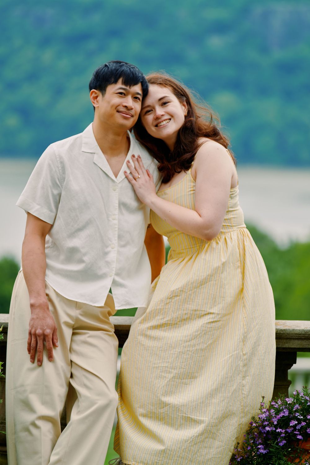 A couple in light summer outfits share a joyful moment outdoors against a scenic lakeside backdrop with mountain views. Wave hill garden