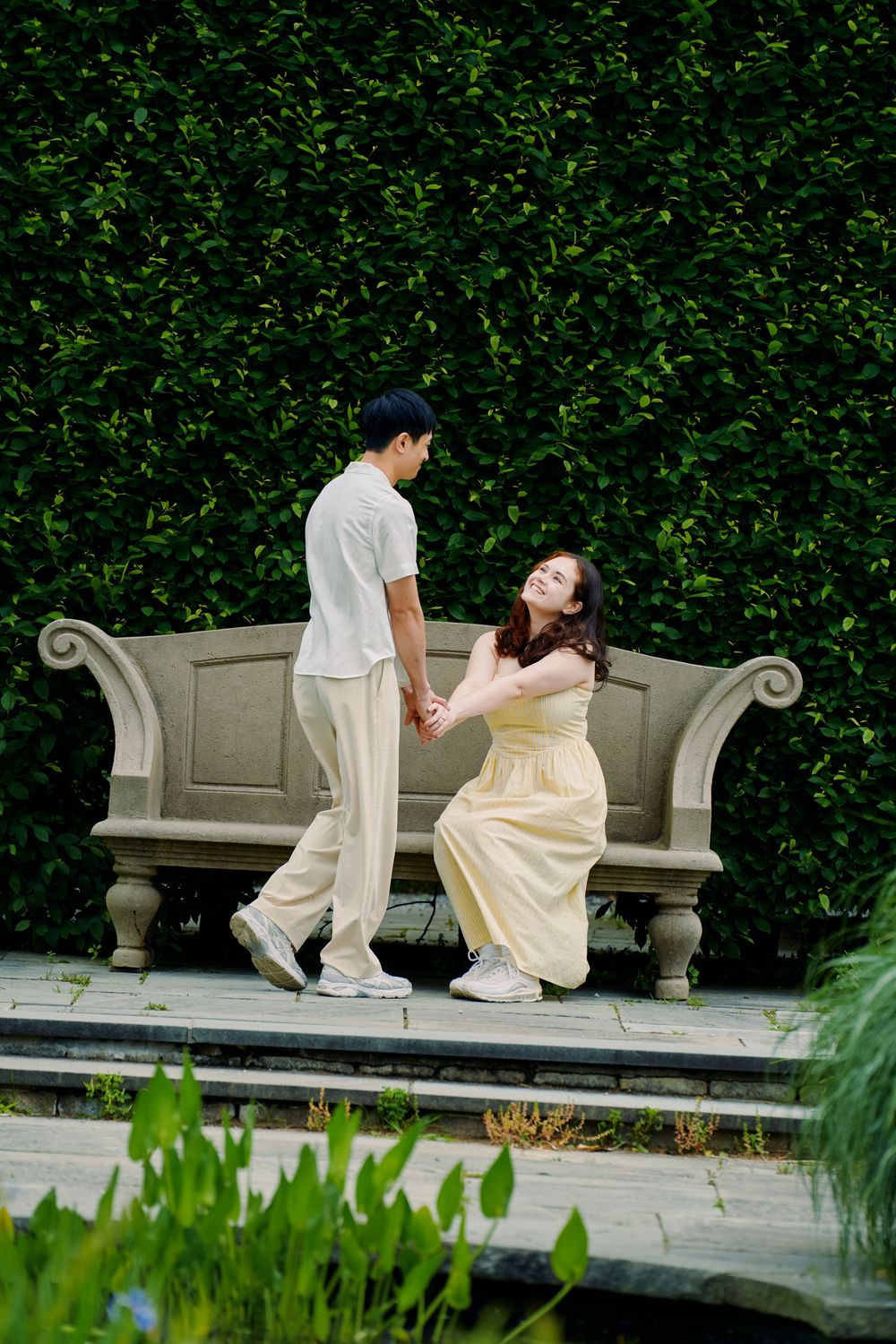 Couple in white outfits sharing a romantic moment on an ornate stone bench against a lush green garden backdrop. Wave hill garden