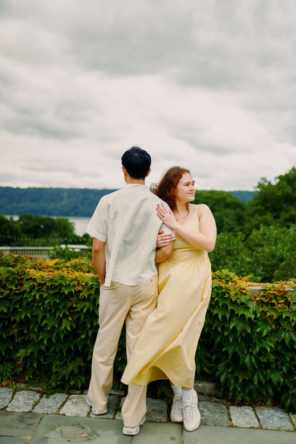 Two people in casual attire sit together on a stone wall overlooking a scenic mountain landscape with greenery. Wave hill garden