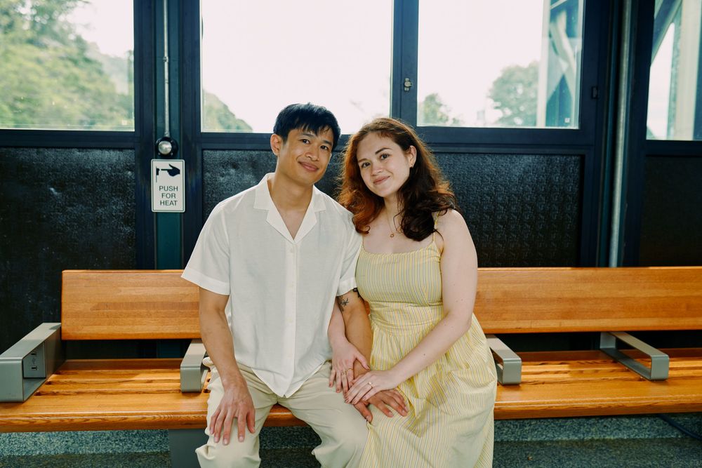 A couple in white and beige outfits pose together on a wooden bench in a bright interior setting. Wave hill garden
