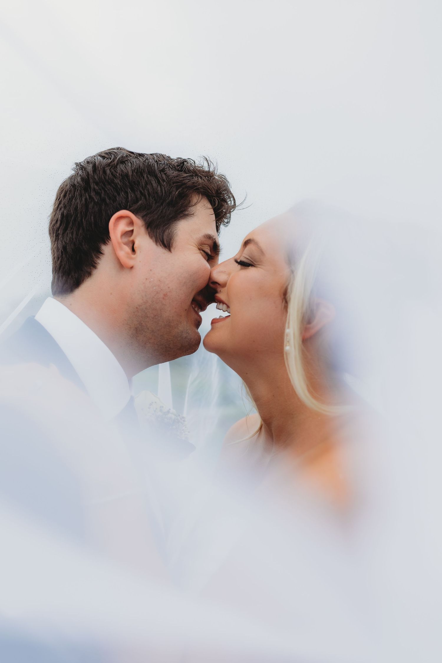 Romantic wedding portrait capturing a tender almost-kiss moment through soft dreamy veil effects.