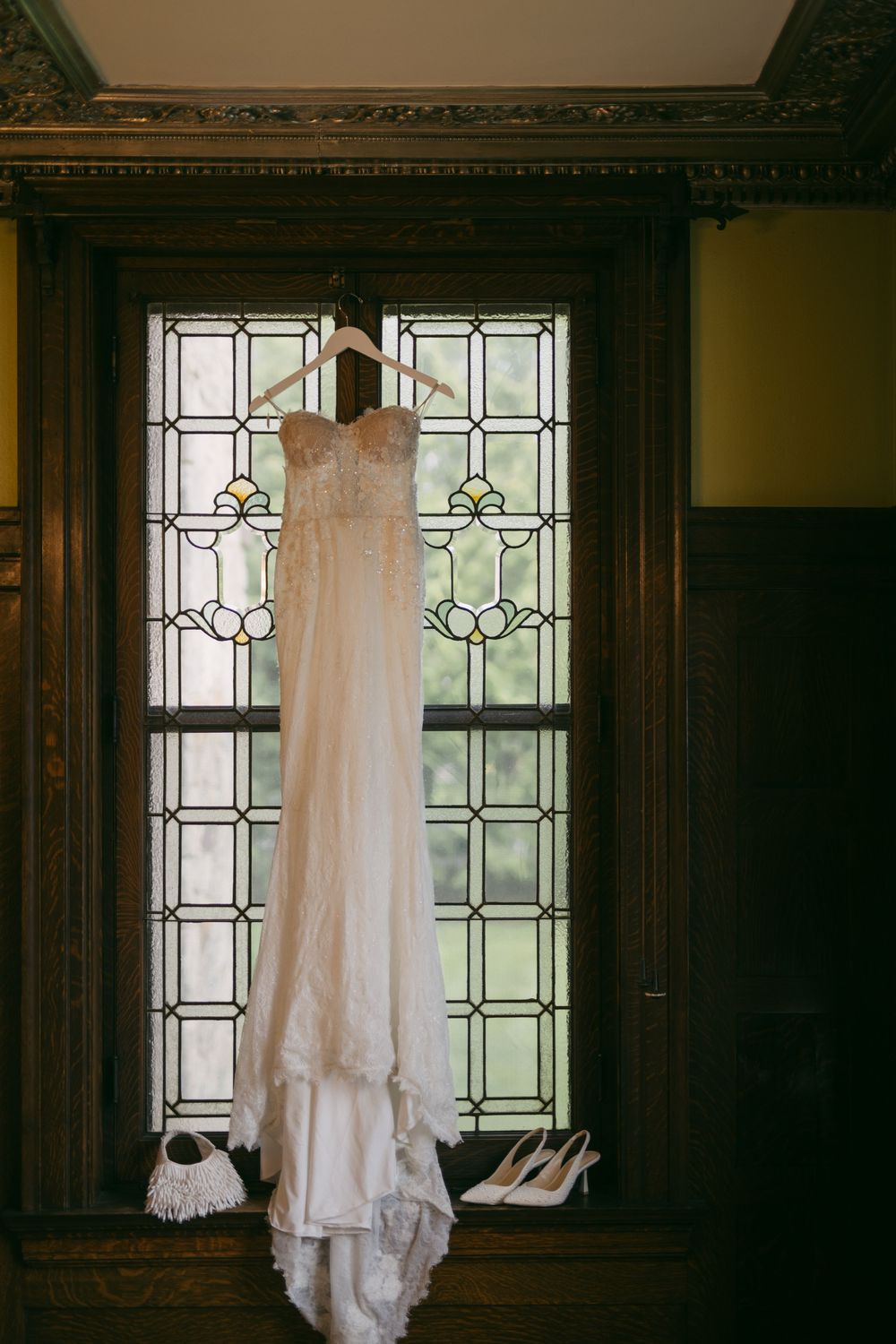 A white lace wedding dress hanging in front of stained glass windows in a dark wood room.