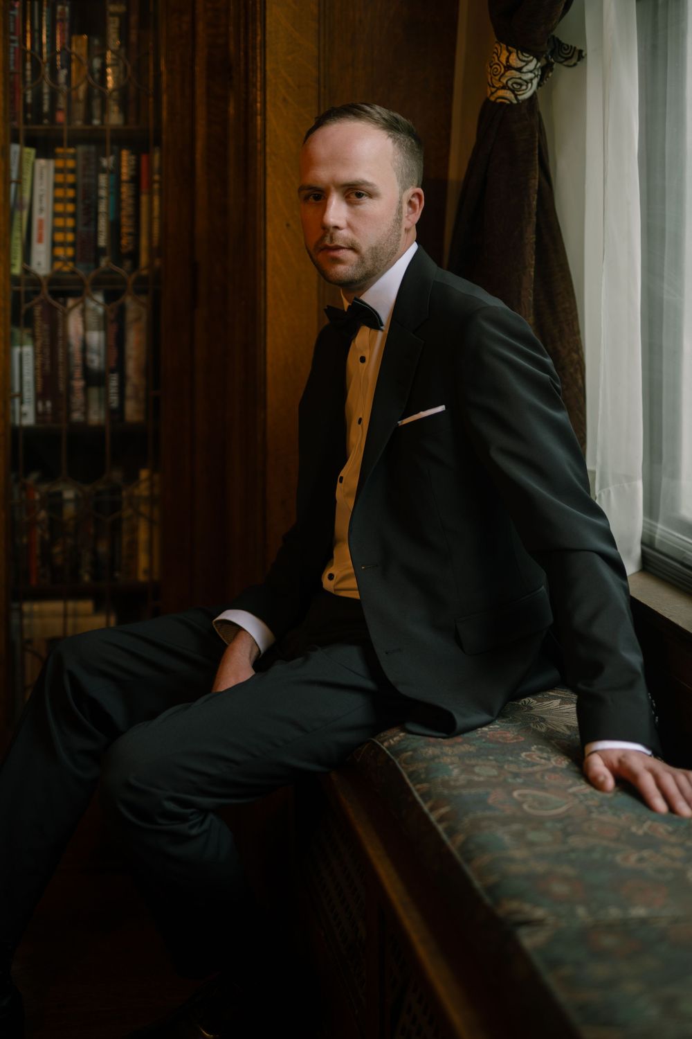 Person in black suit sitting on upholstered bench in wood-paneled room.