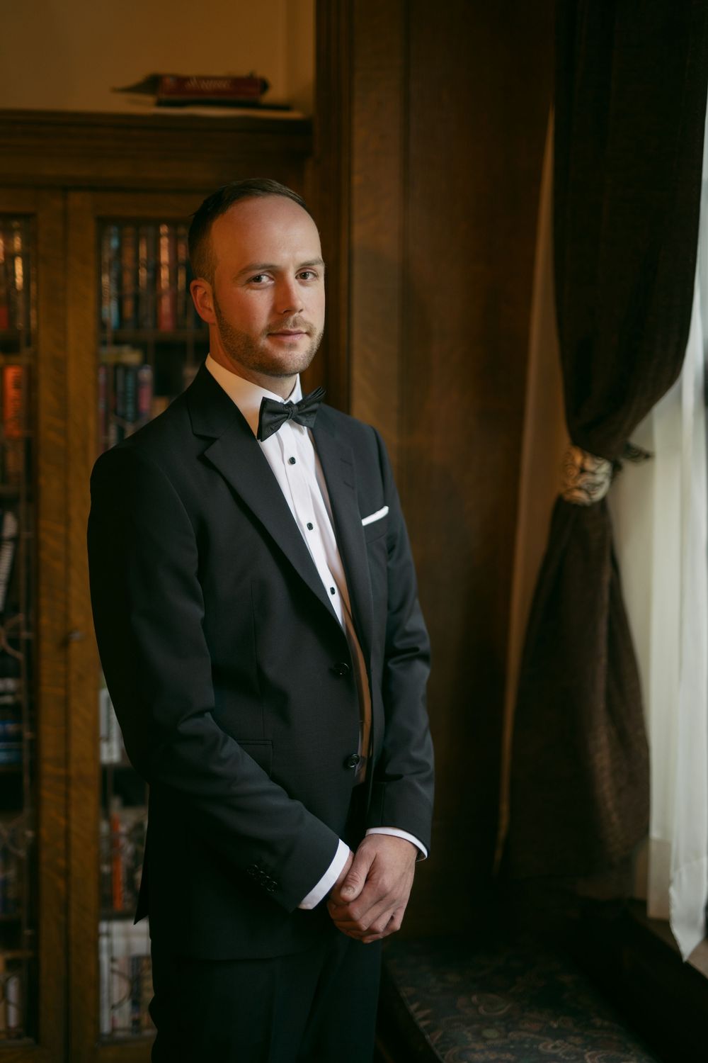 Person wearing black tuxedo standing in elegant wood-paneled room.