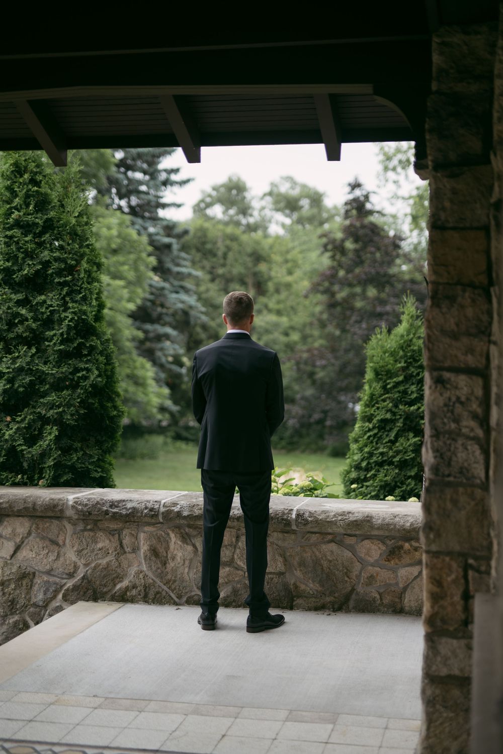Person in dark suit standing on stone patio overlooking green landscape.