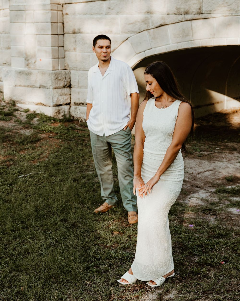 A couple in wedding attire walking together in front of a rustic stone wall and grass background.