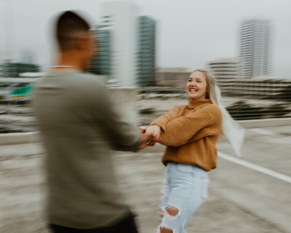 A couple dances and spins together on a rooftop with city buildings visible in the background.