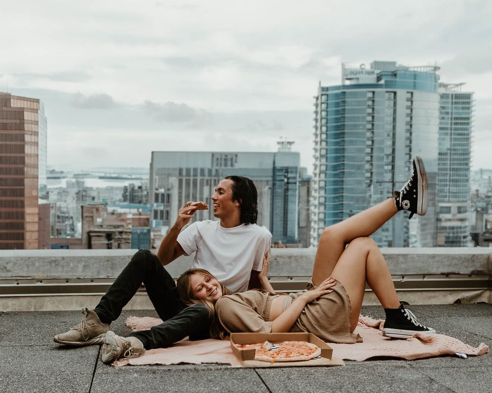 Two people lying down and laughing together on a rooftop with city buildings in the background.