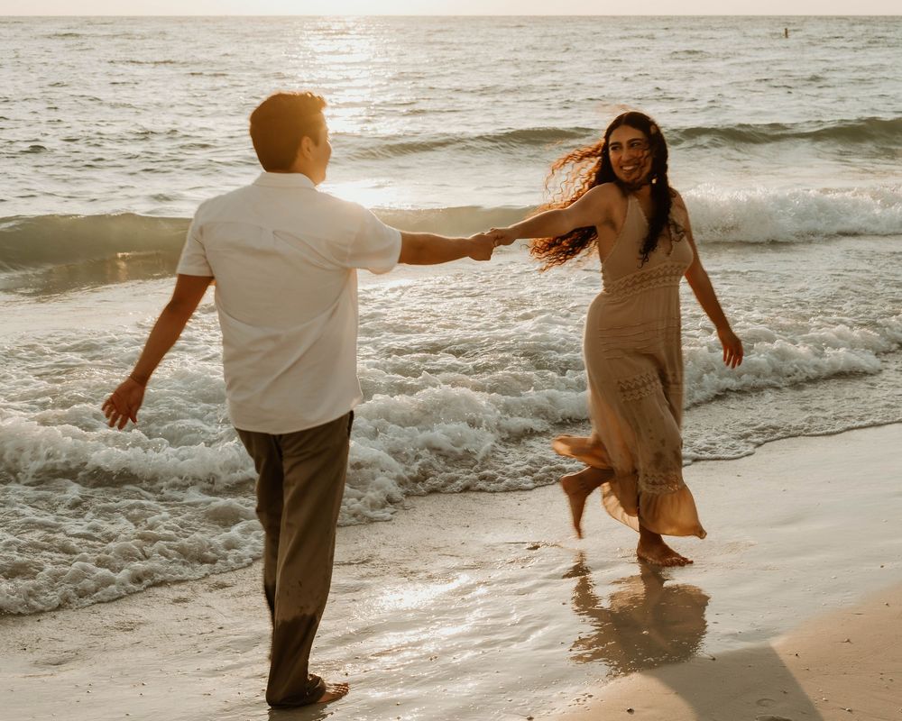 A joyful couple dances together on a sandy beach at sunset with waves rolling in the background.
