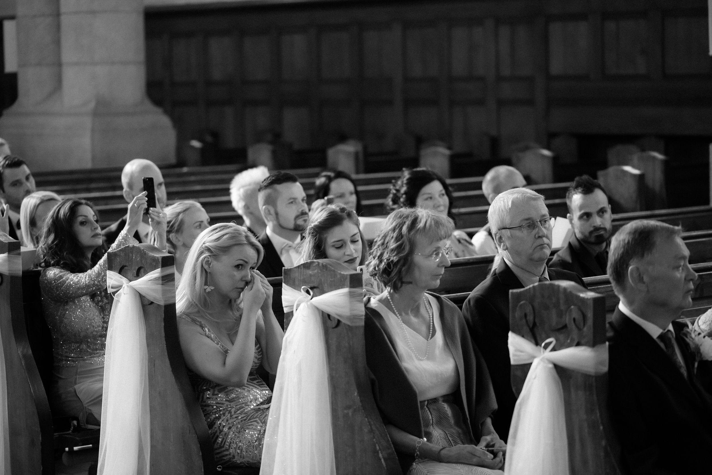 Black and white photo of wedding ceremony guests seated in decorated church pews.