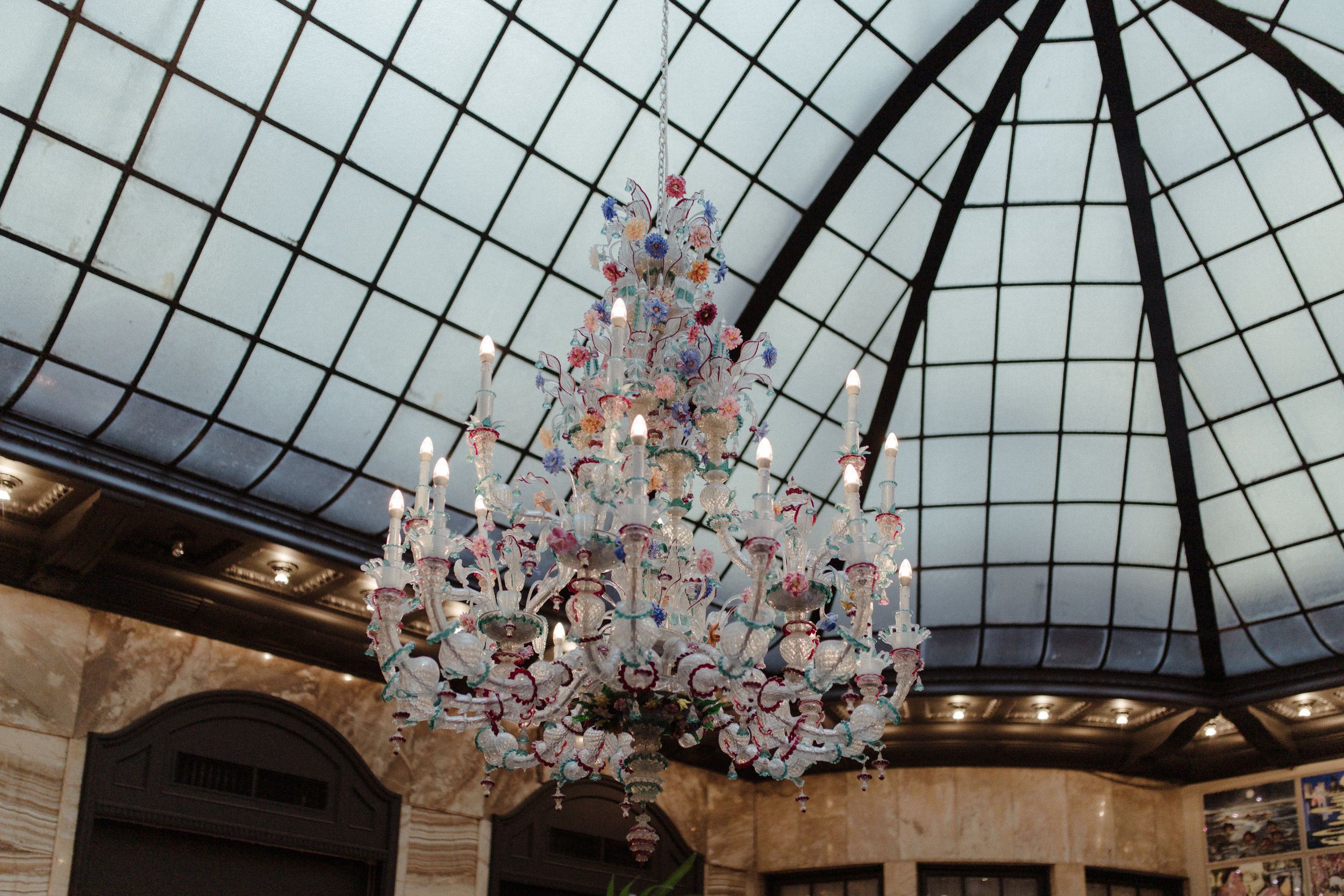 Ornate crystal chandelier hanging from a glass-paneled domed ceiling in an elegant venue.
