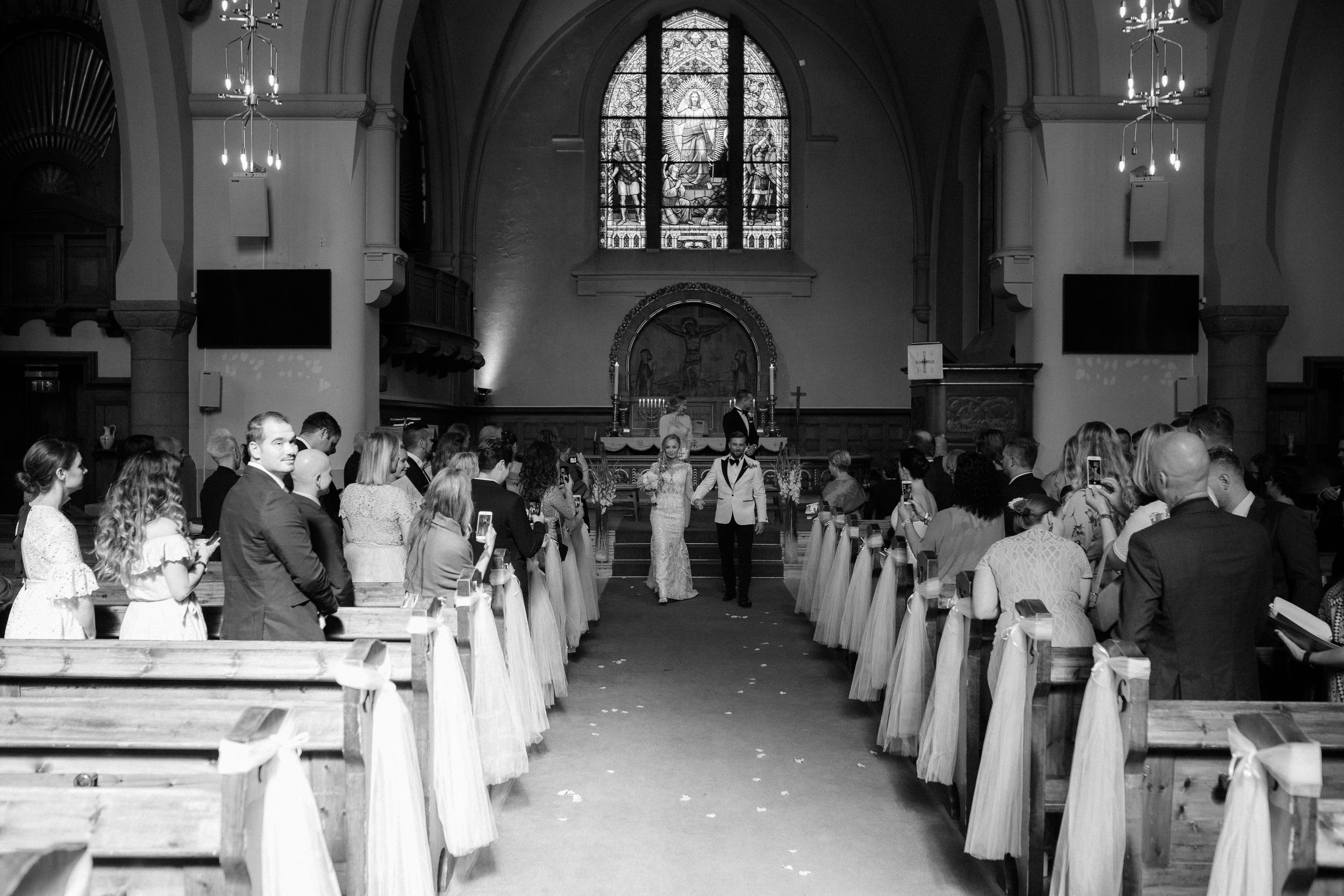 Black and white panorama of an elegant church interior with rows of white pews and arched windows.