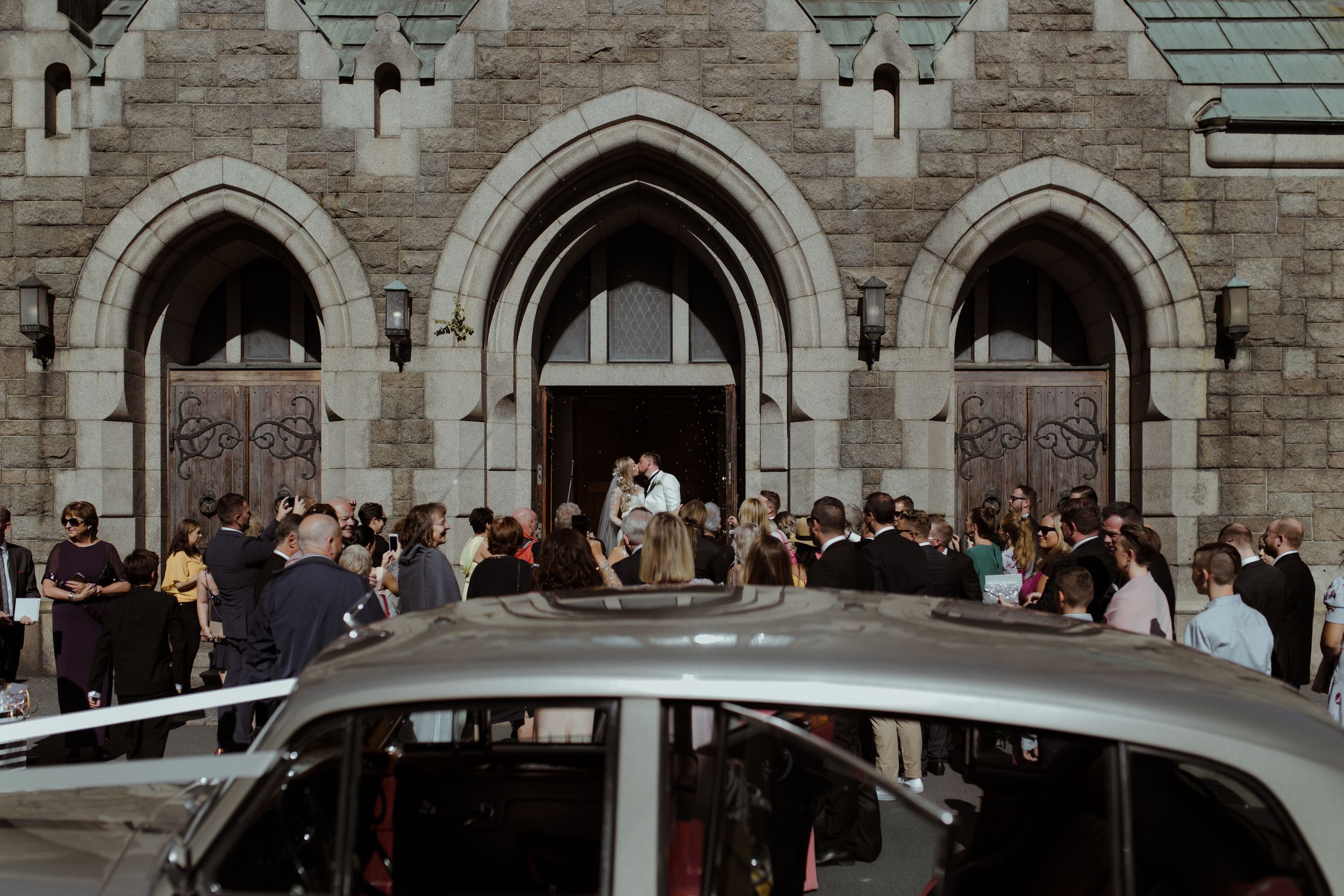 Stone church exterior with Gothic arched windows and gathering crowd outside.