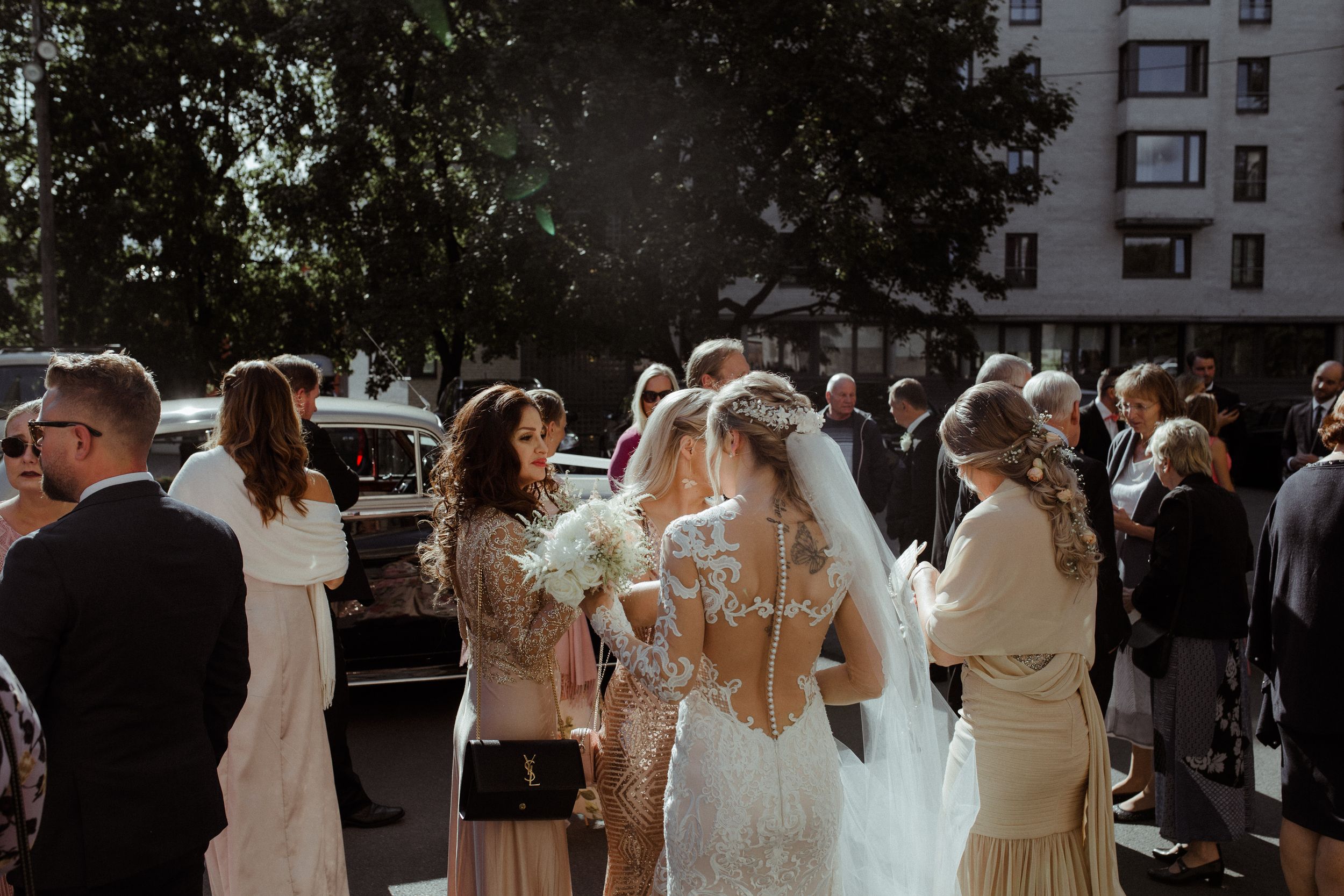 Wedding guests gather outside on a sunny day with elegant attire and flowing dresses.