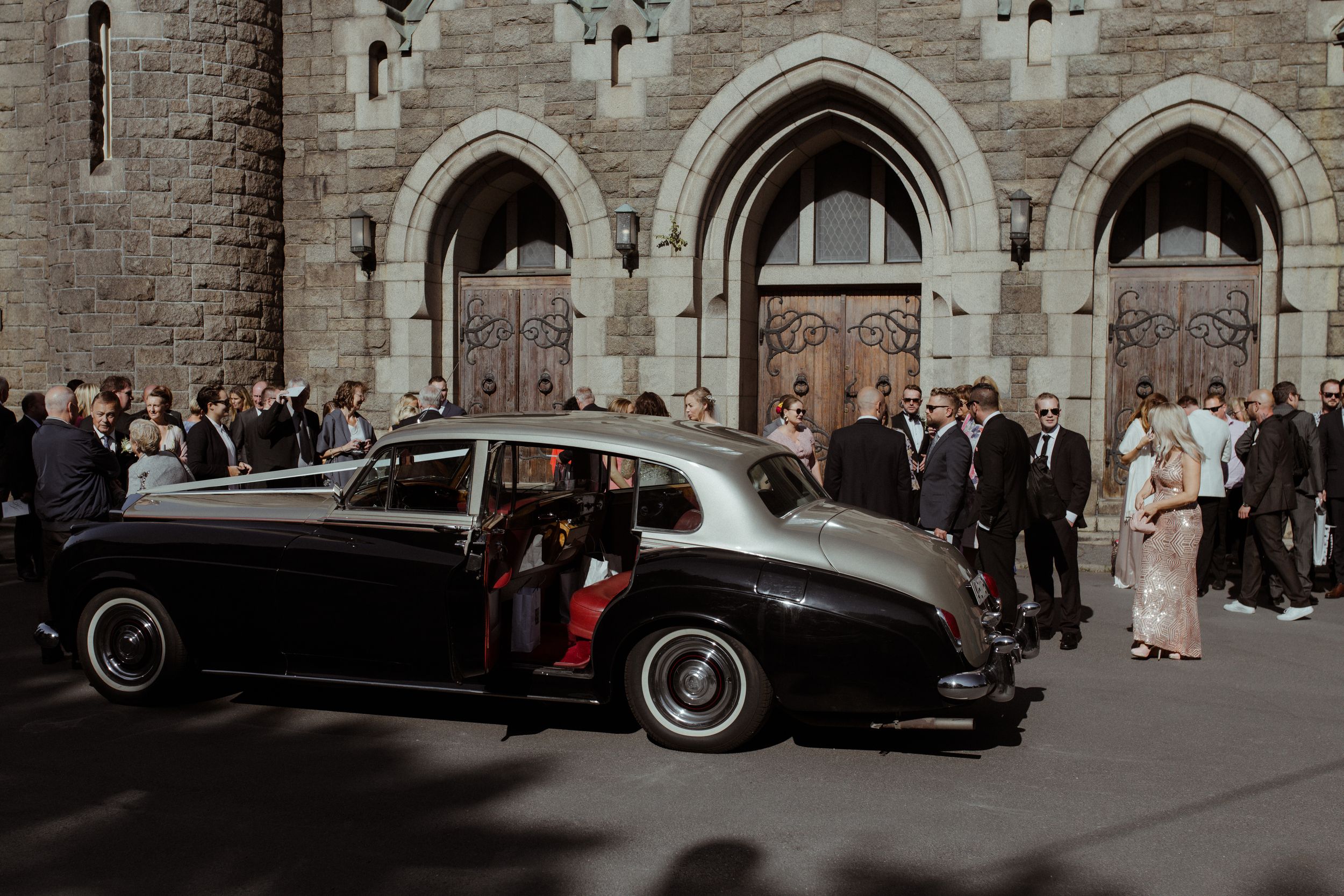 Vintage cars parked outside stone church building with wedding guests gathering on sidewalk.