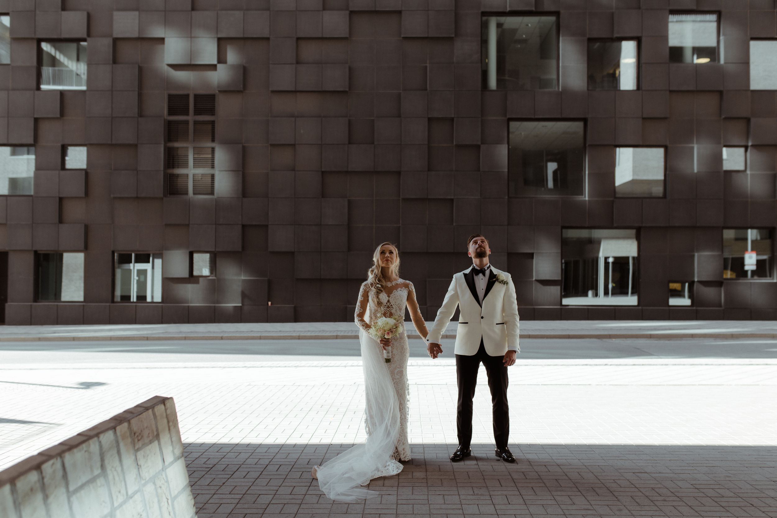 Couple stands against modern architectural backdrop with geometric patterns and windows.