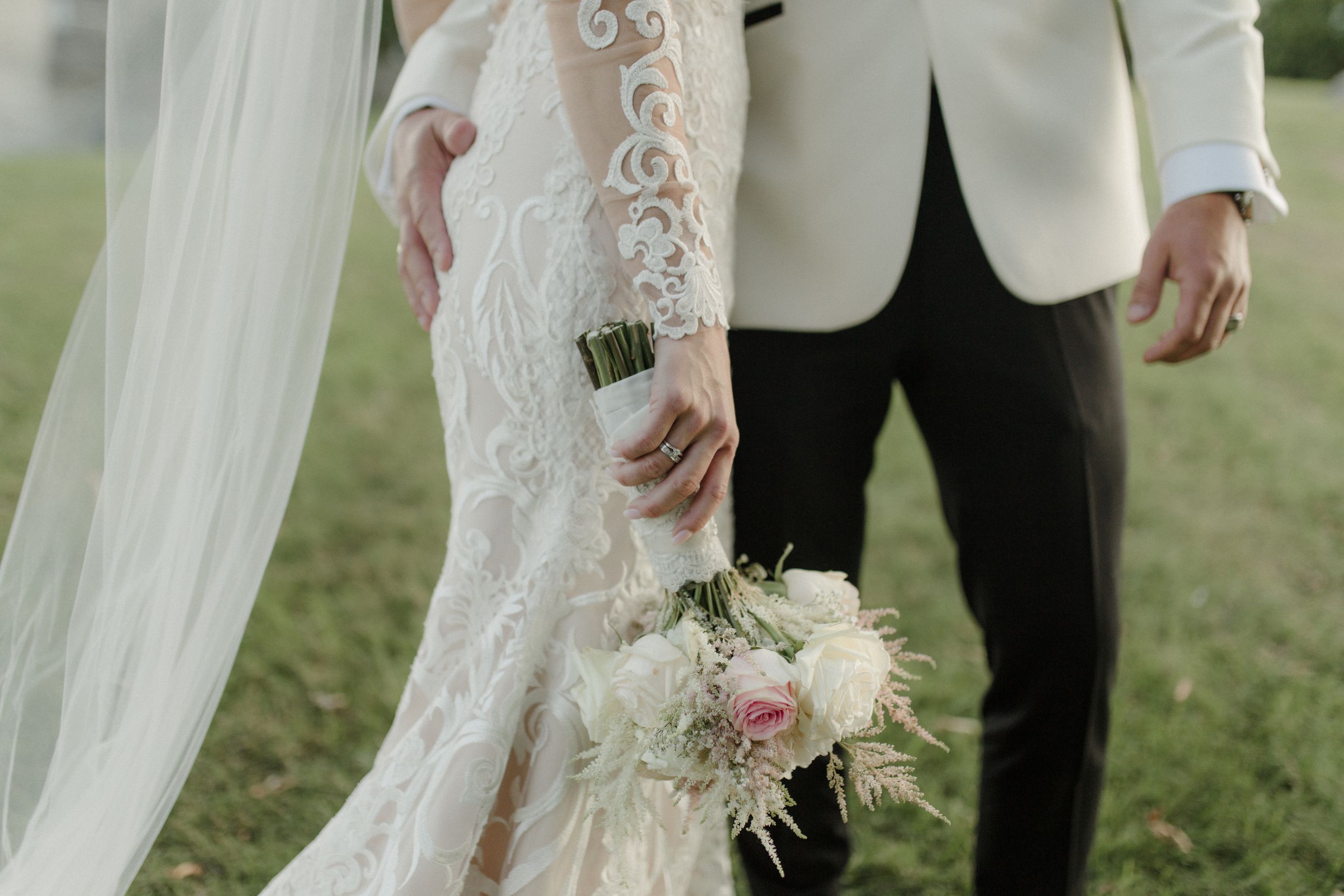 Close-up detail of lace wedding dress sleeve and flowing veil against green grass.