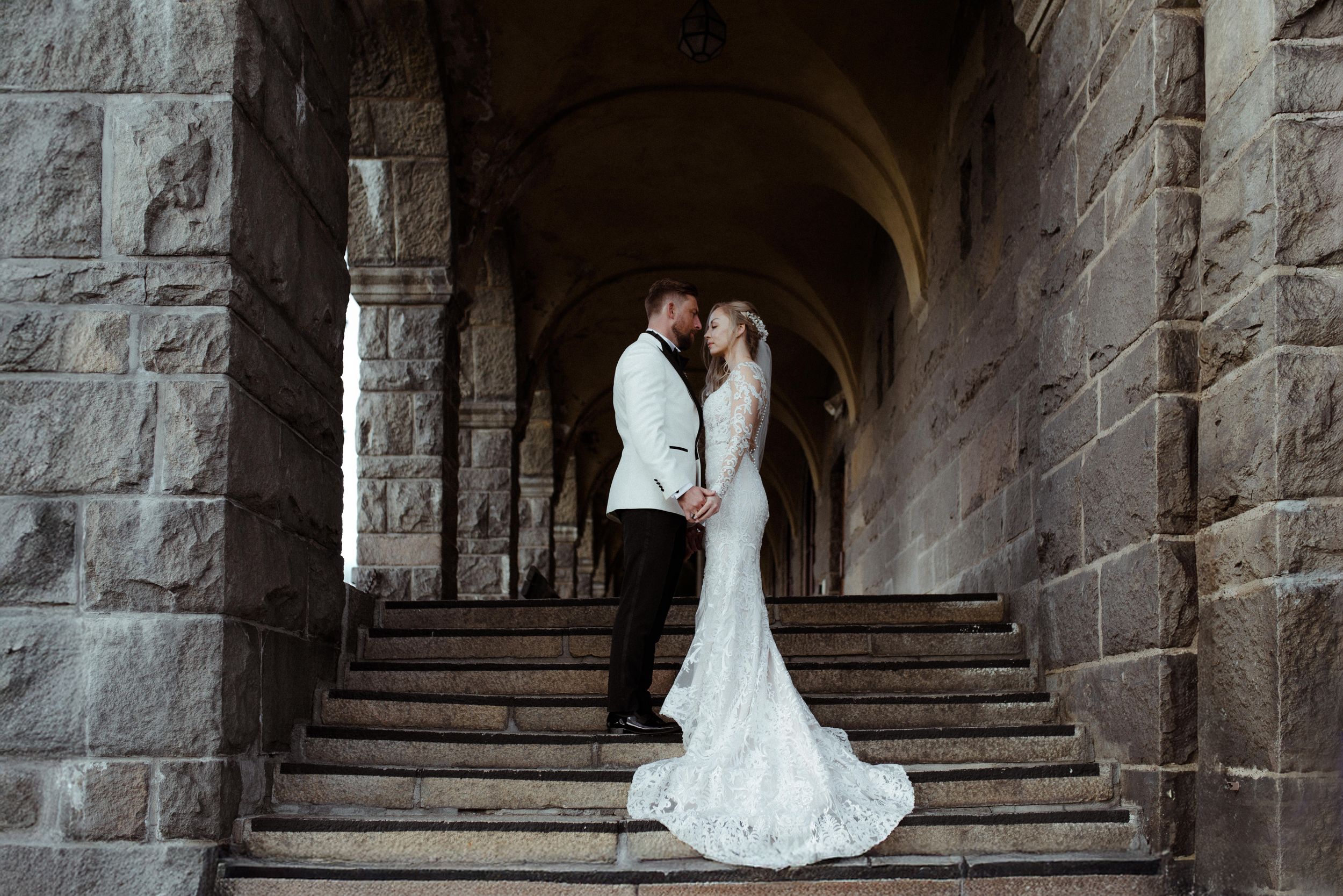 A wedding couple stands in a dramatic stone archway with steps in an architectural setting.