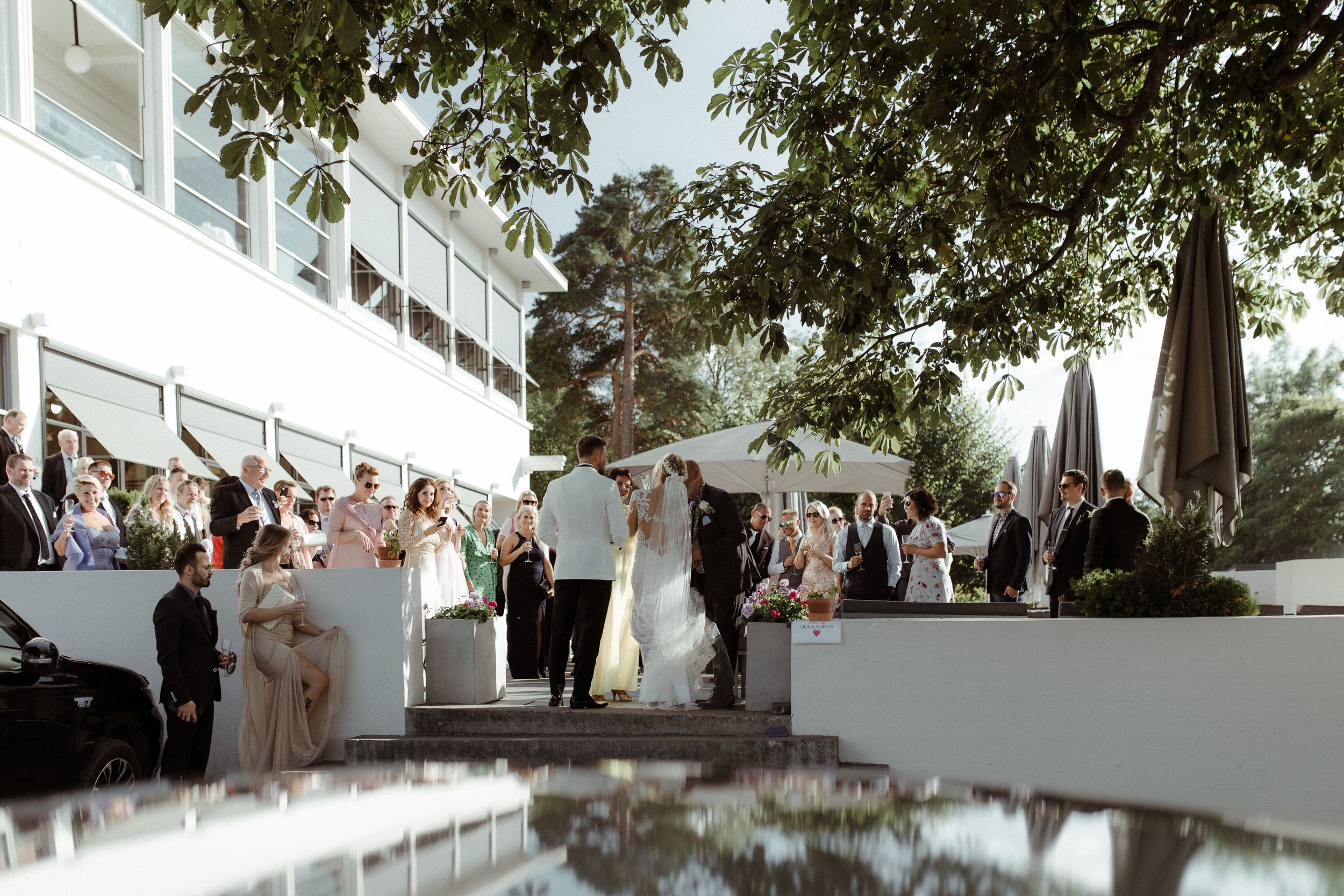Outdoor wedding ceremony and reception with guests gathered around a reflective pool during sunset.