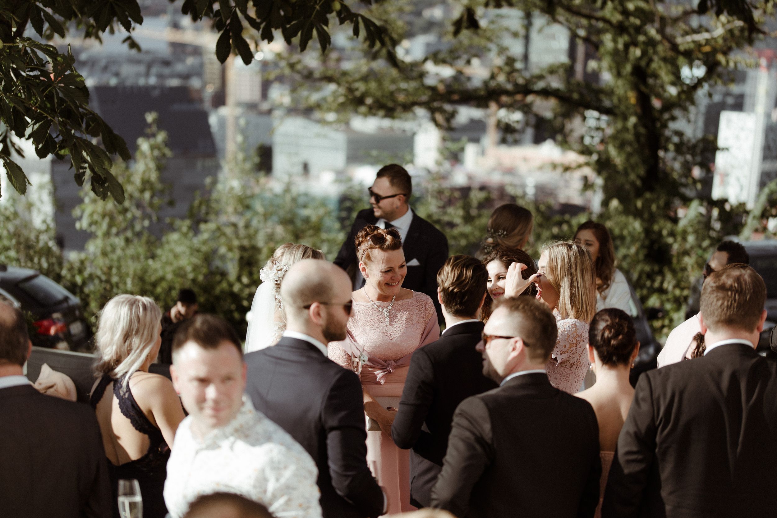 A group of wedding guests gather at an elegant outdoor reception under trees and greenery.